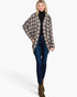 A woman modeling a beige and dark gray houndstooth patterned open-front cardigan over a black turtleneck, paired with dark blue skinny jeans and brown ankle boots, against a plain white studio background.