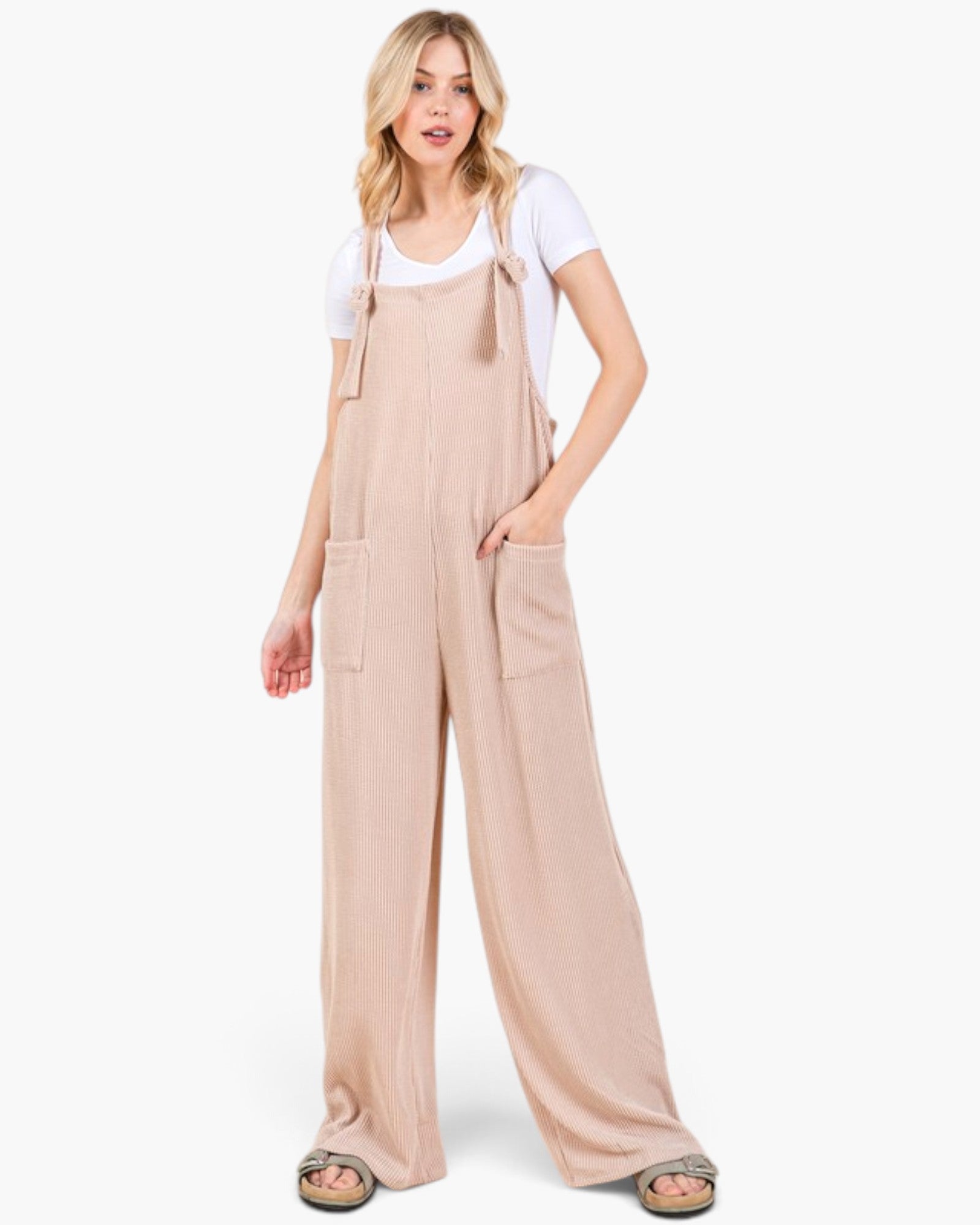 A woman modeling wide-leg beige overall jumpsuit over a white t-shirt, standing against a plain white studio background.