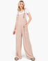 A woman modeling wide-leg beige overall jumpsuit over a white t-shirt, standing against a plain white studio background.