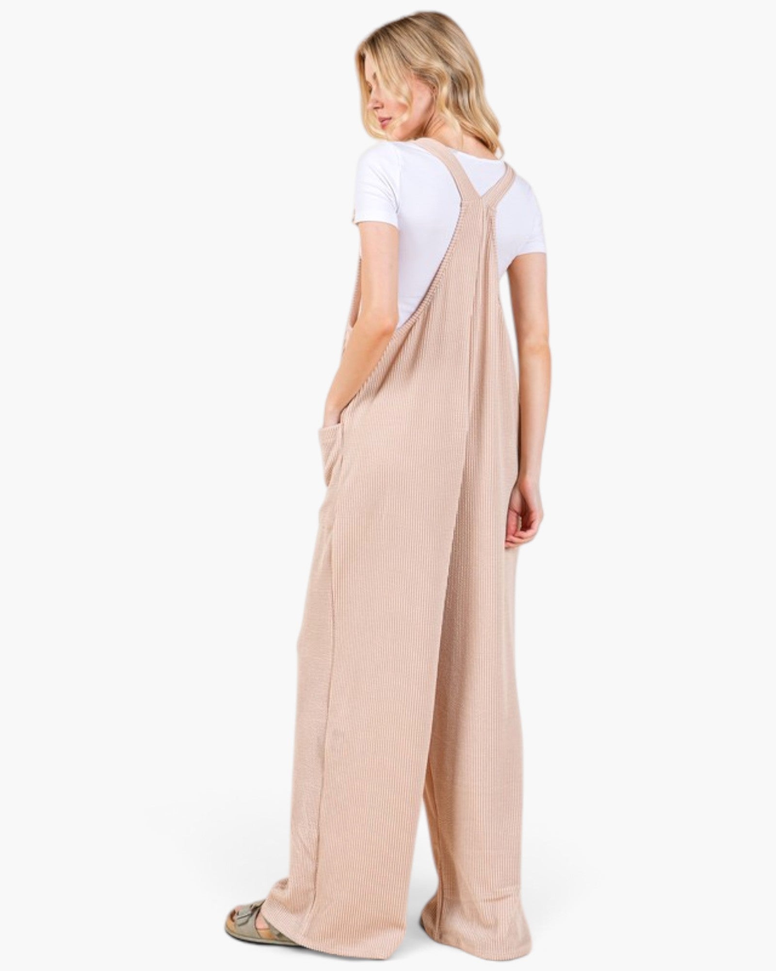 A woman shown from the back wearing a light beige wide-leg overall jumpsuit over a white short-sleeve t-shirt, photographed against a clean white studio background.