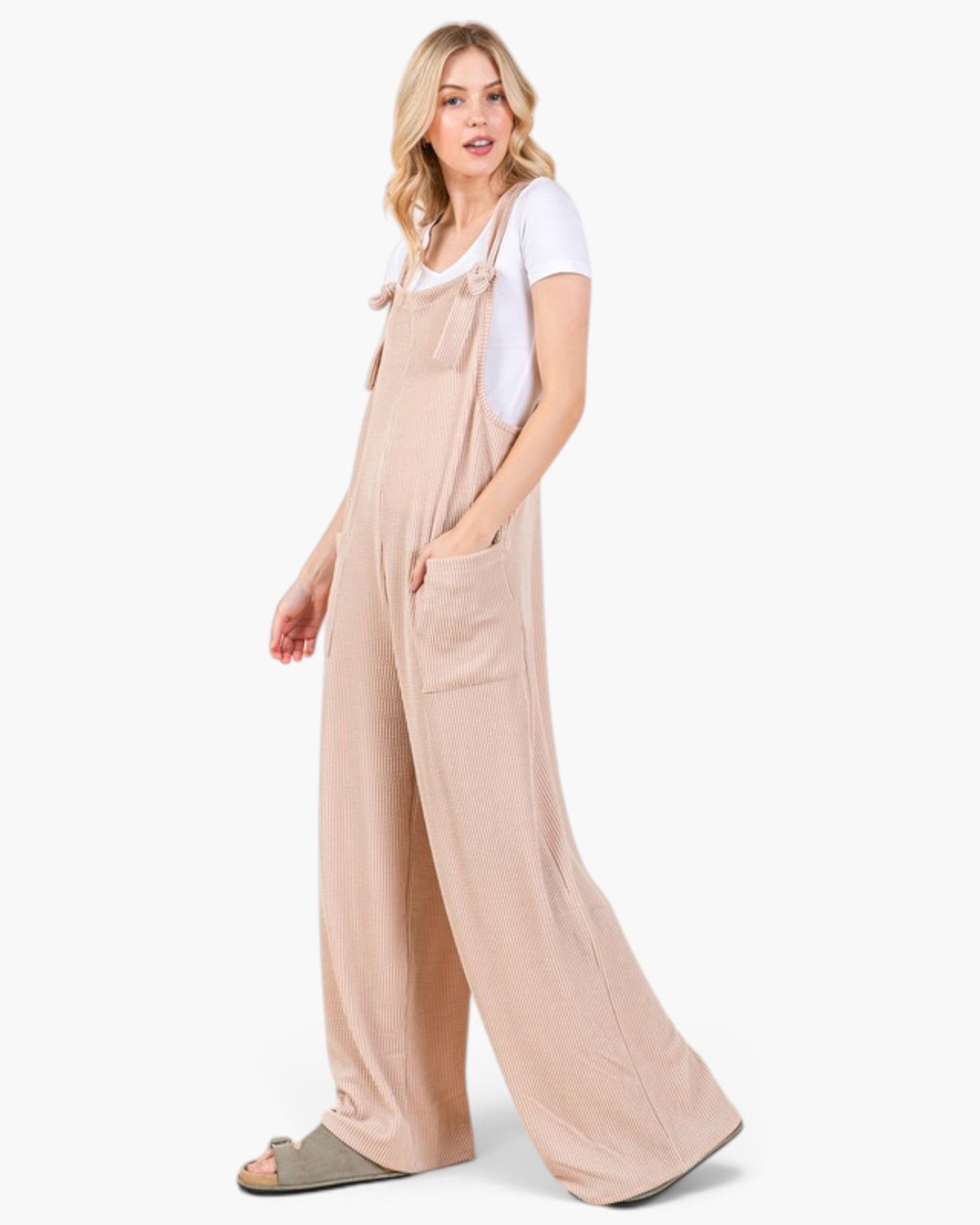 A woman modeling wide-leg beige overall jumpsuit with a white t-shirt underneath and gray slide sandals, standing against a plain white studio background.