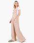 A woman modeling wide-leg beige overall jumpsuit with a white t-shirt underneath and gray slide sandals, standing against a plain white studio background.