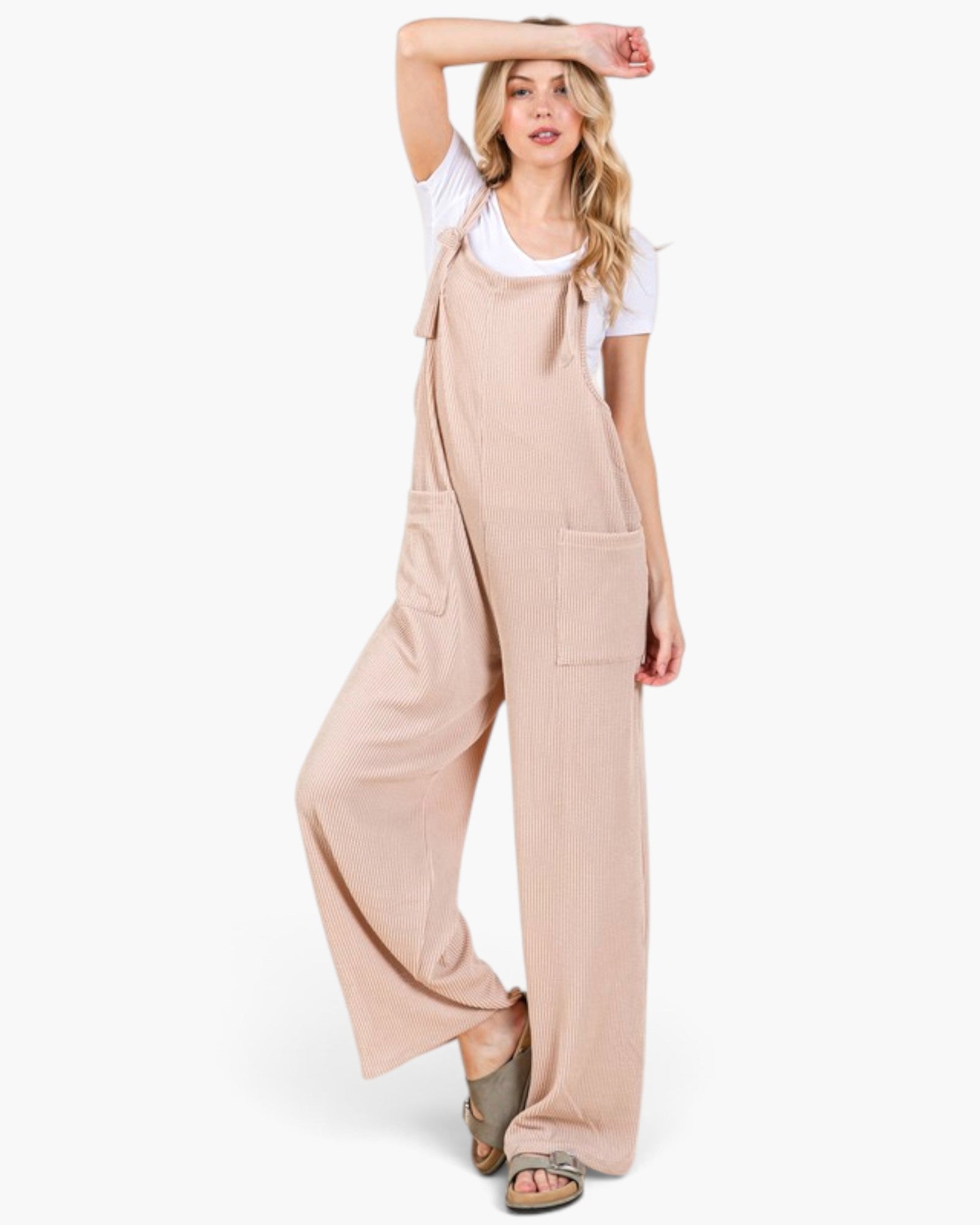A woman modeling a light beige wide-leg jumpsuit with front patch pockets over a white t-shirt and brown sandals, set against a plain white studio background.