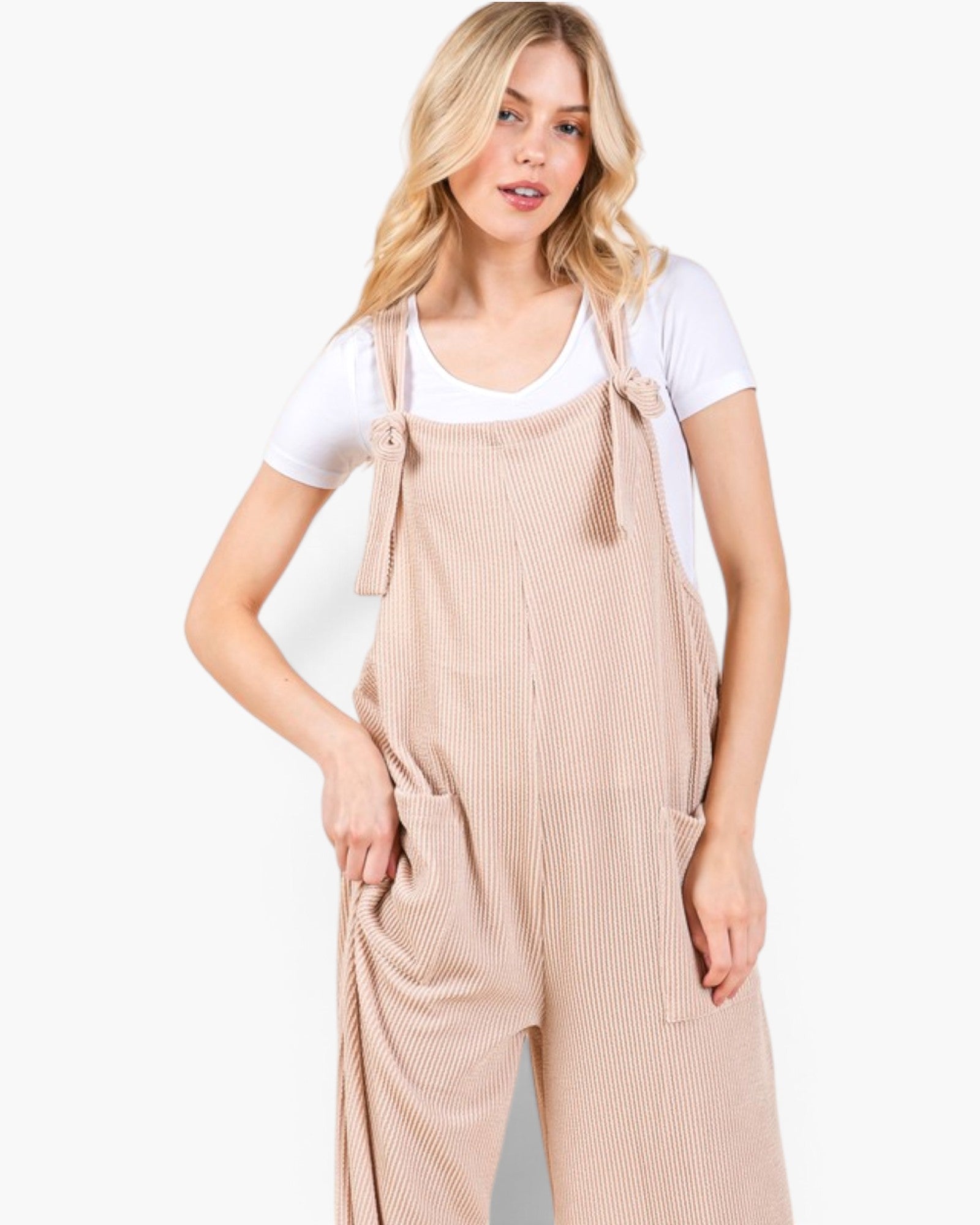 A woman modeling light beige corduroy bib overalls with tie straps over a white t-shirt, photographed against a clean white studio background.