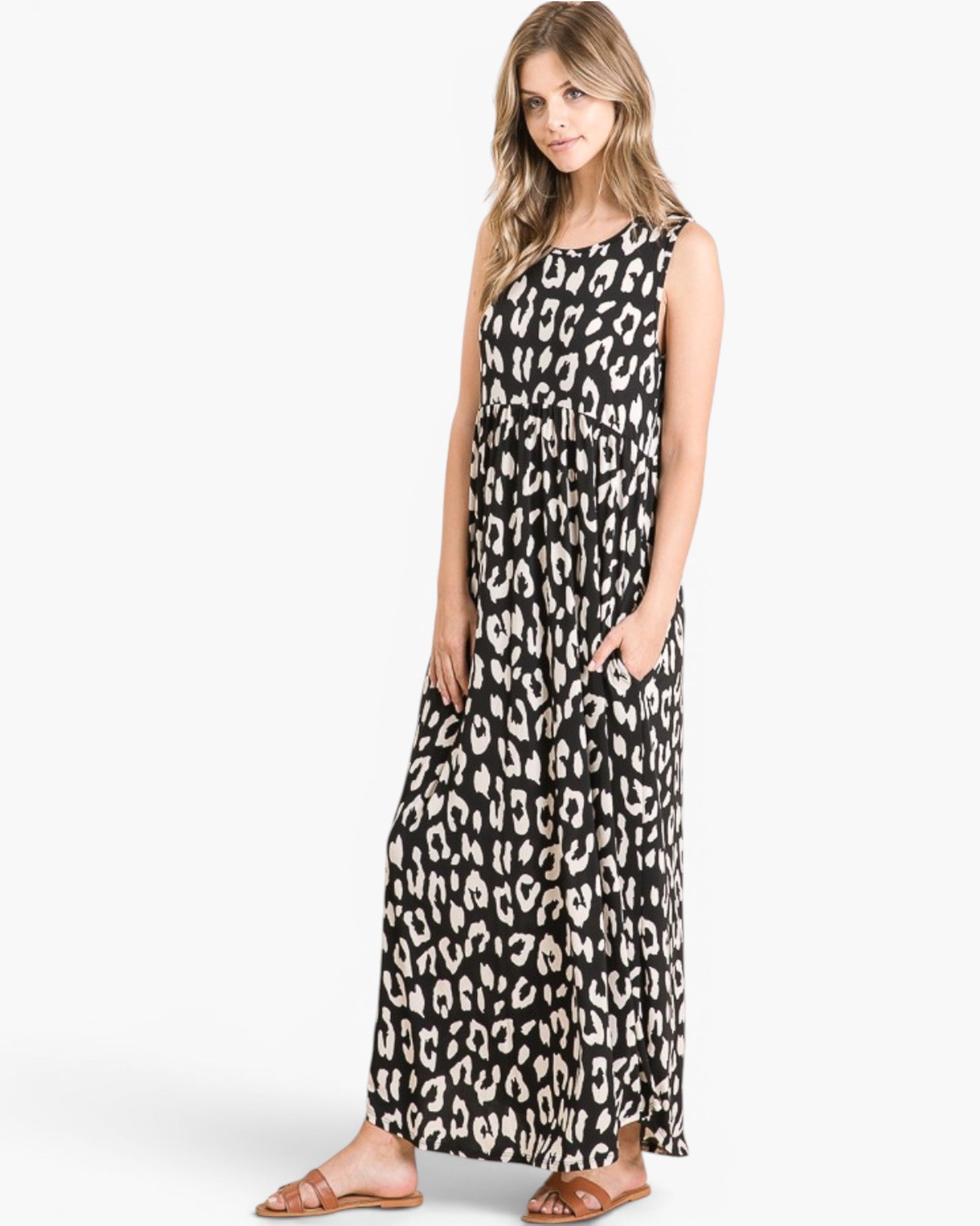 A woman modeling a sleeveless black and white leopard-print maxi dress with side pockets, paired with brown flat sandals, against a light gray studio background.