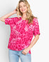 A woman wearing a bright pink short-sleeve top with a darker pink leopard print pattern, paired with light blue jeans, against a plain light gray background.