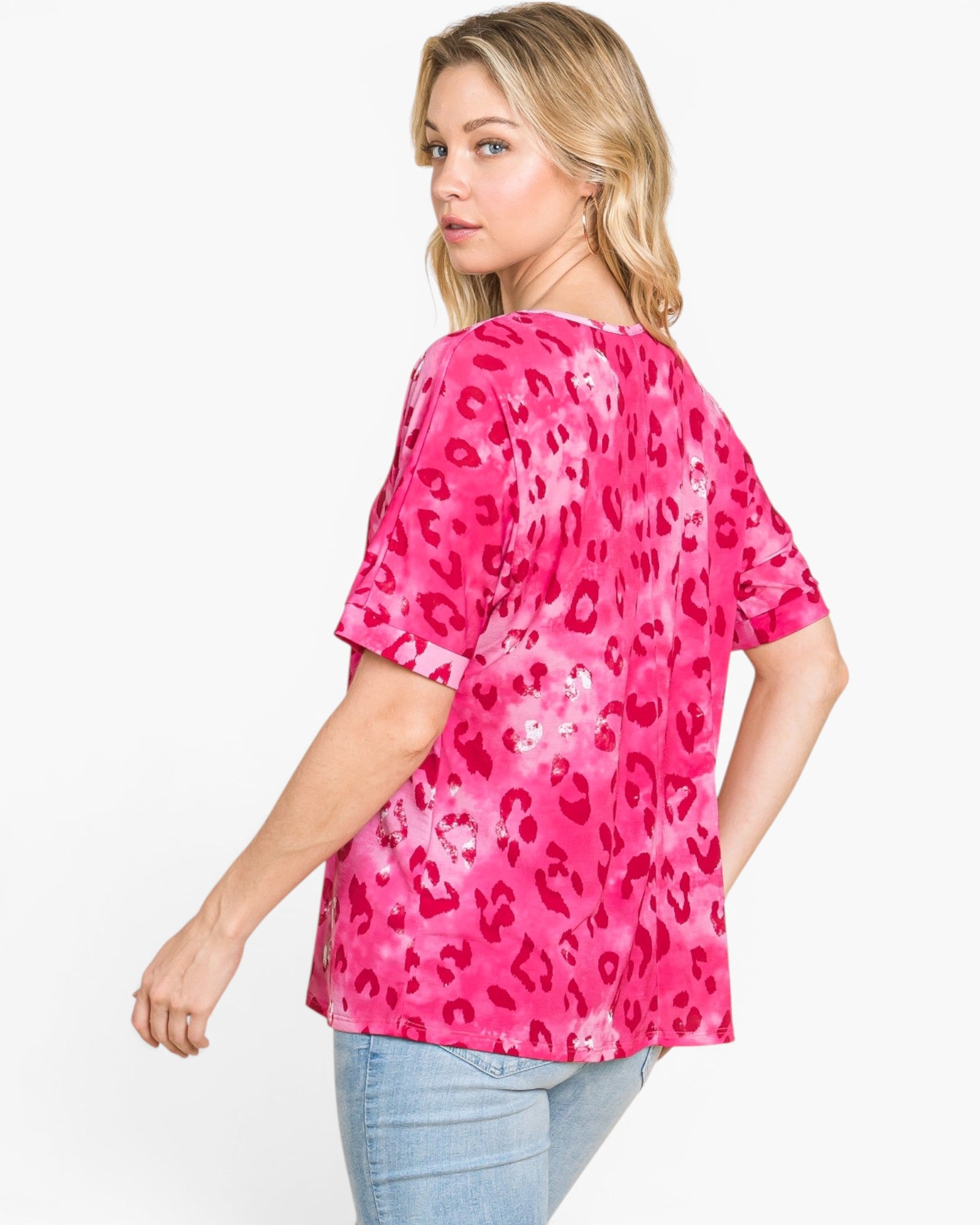A woman wearing a bright pink short-sleeve top with a darker pink leopard print pattern, paired with light blue jeans, photographed against a plain white studio background.
