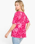 A woman wearing a bright pink short-sleeve top with a darker pink leopard print pattern, paired with light blue jeans, photographed against a plain white studio background.