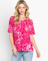 A woman modeling a short-sleeve pink top with a darker pink leopard print pattern, paired with light blue jeans, against a plain white background.