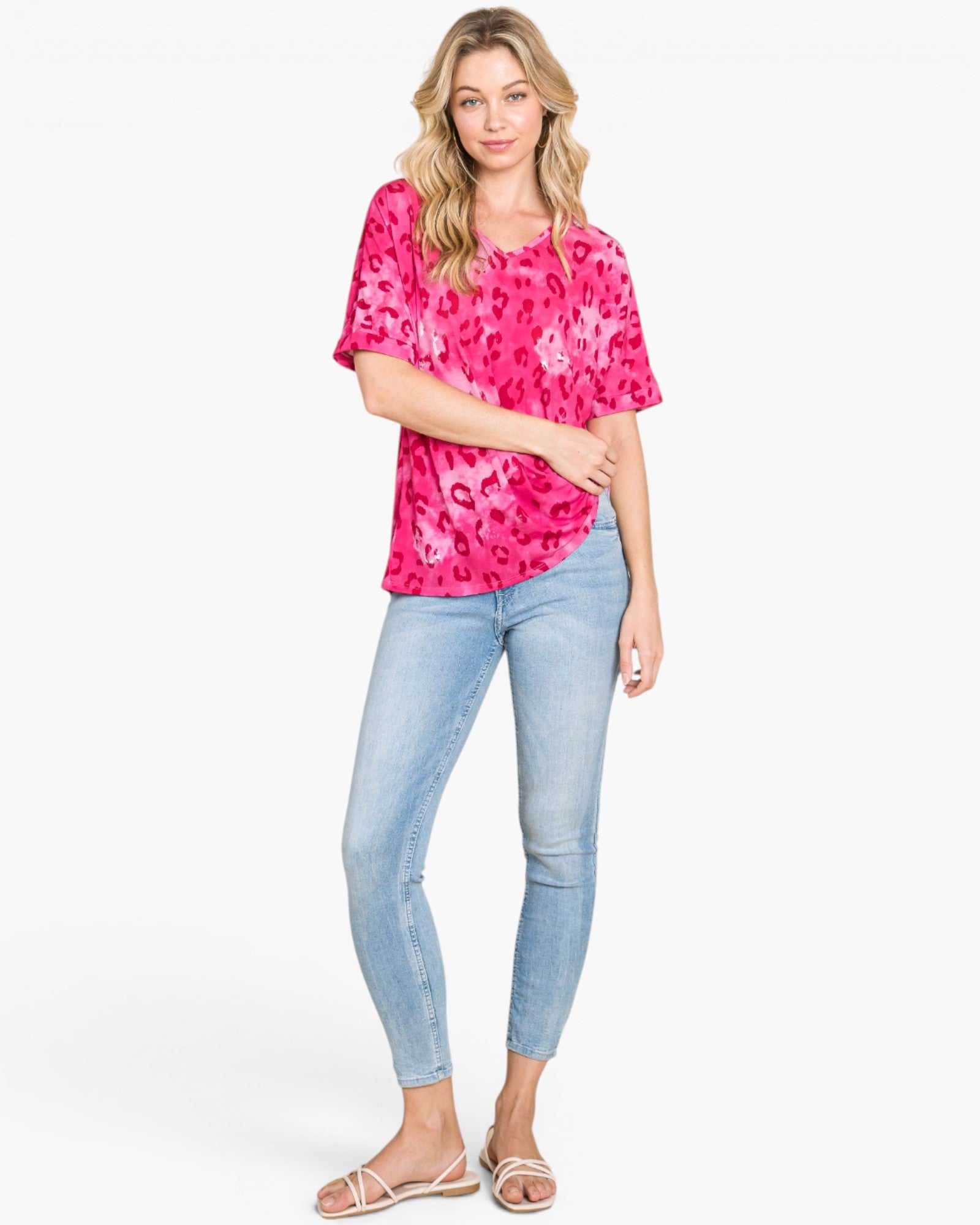 A woman modeling a bright pink short-sleeve top with a tonal leopard print, paired with light-wash blue skinny jeans and beige flat sandals, against a plain white studio background.