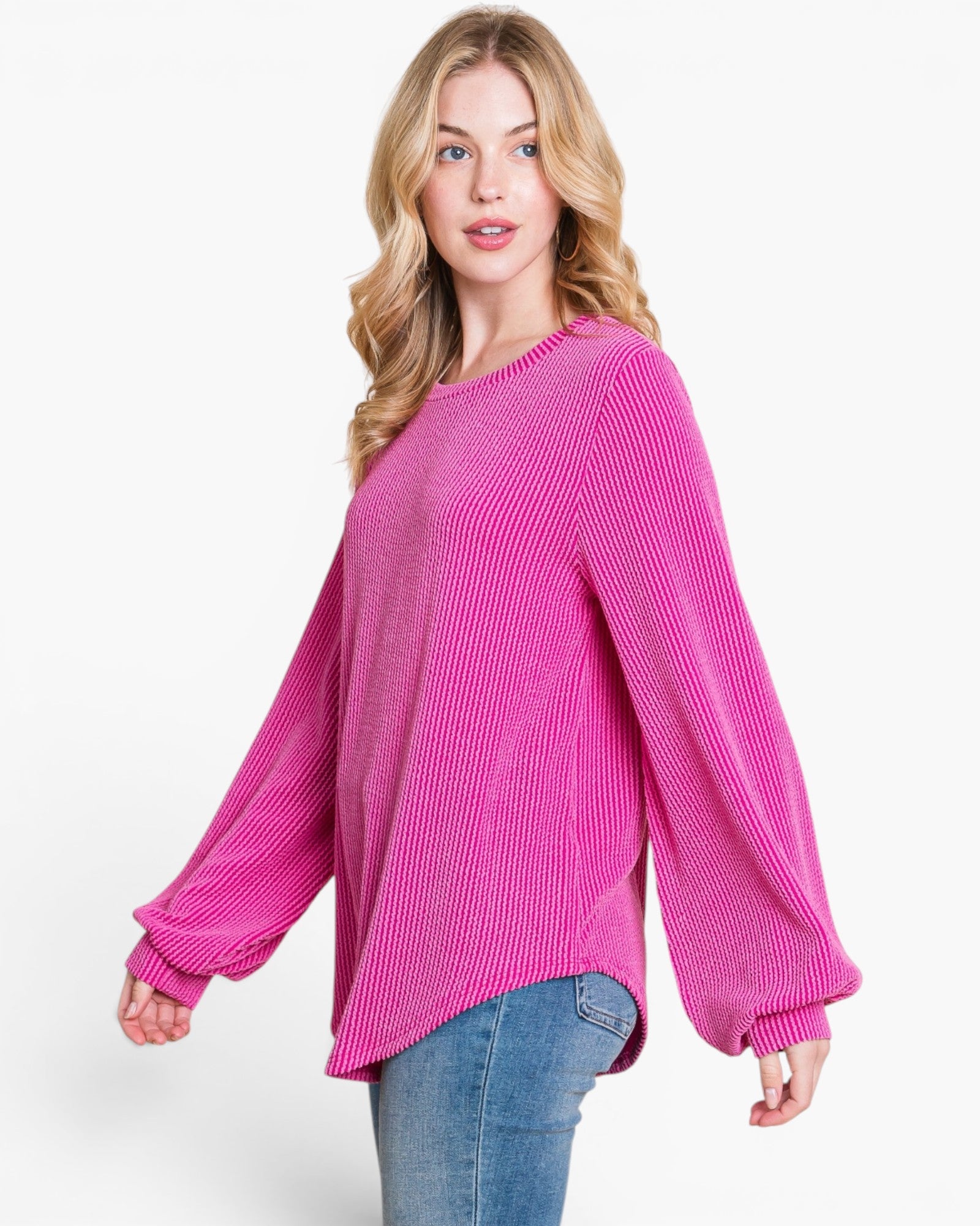A woman modeling a bright pink ribbed knit sweater with balloon sleeves and a relaxed fit, paired with blue jeans against a plain white studio background.