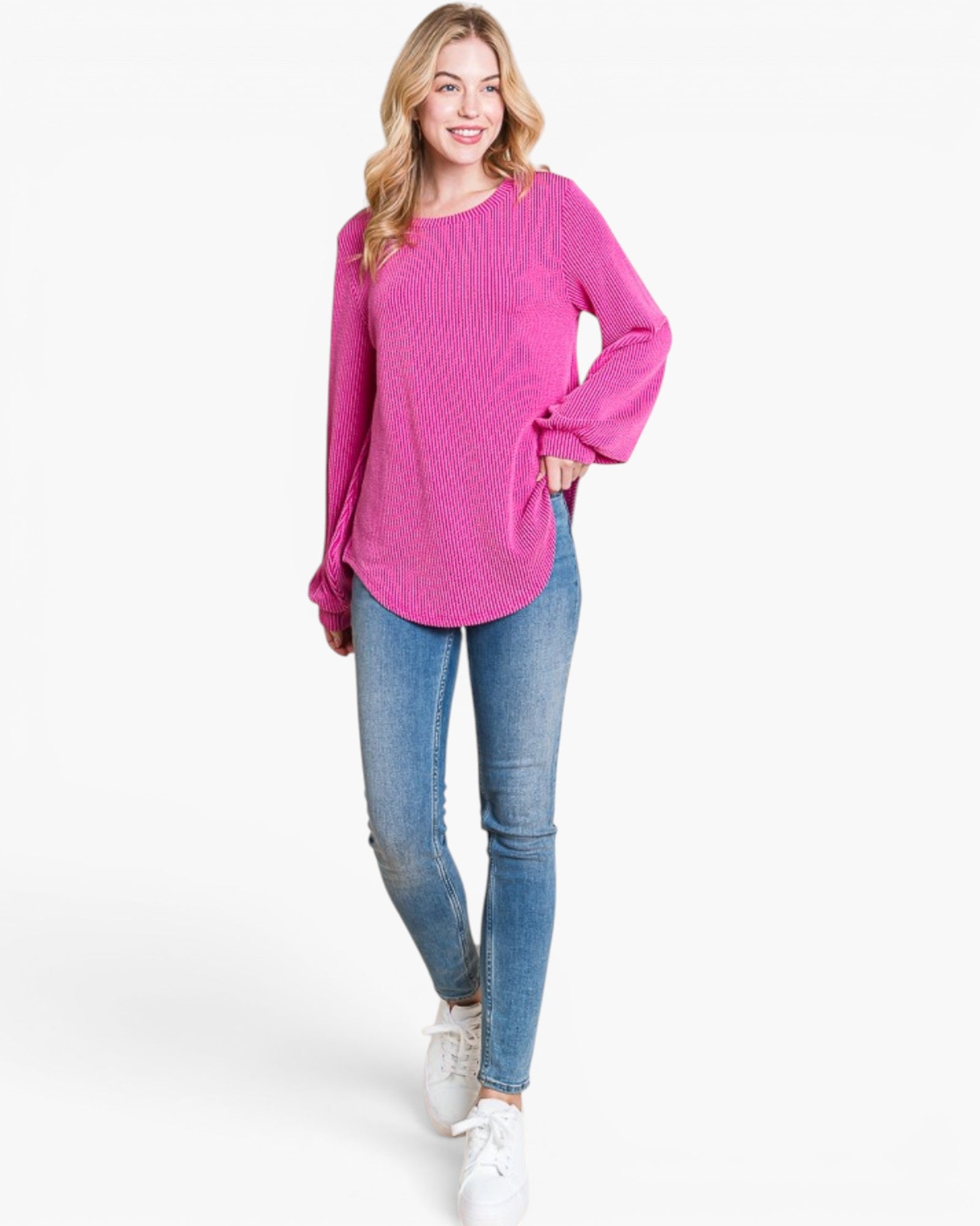 A woman modeling a bright pink long-sleeve knit top with flared cuffs, paired with blue skinny jeans and white sneakers, against a plain white studio background.