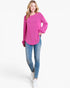 A woman modeling a bright pink long-sleeve knit top with flared cuffs, paired with blue skinny jeans and white sneakers, against a plain white studio background.