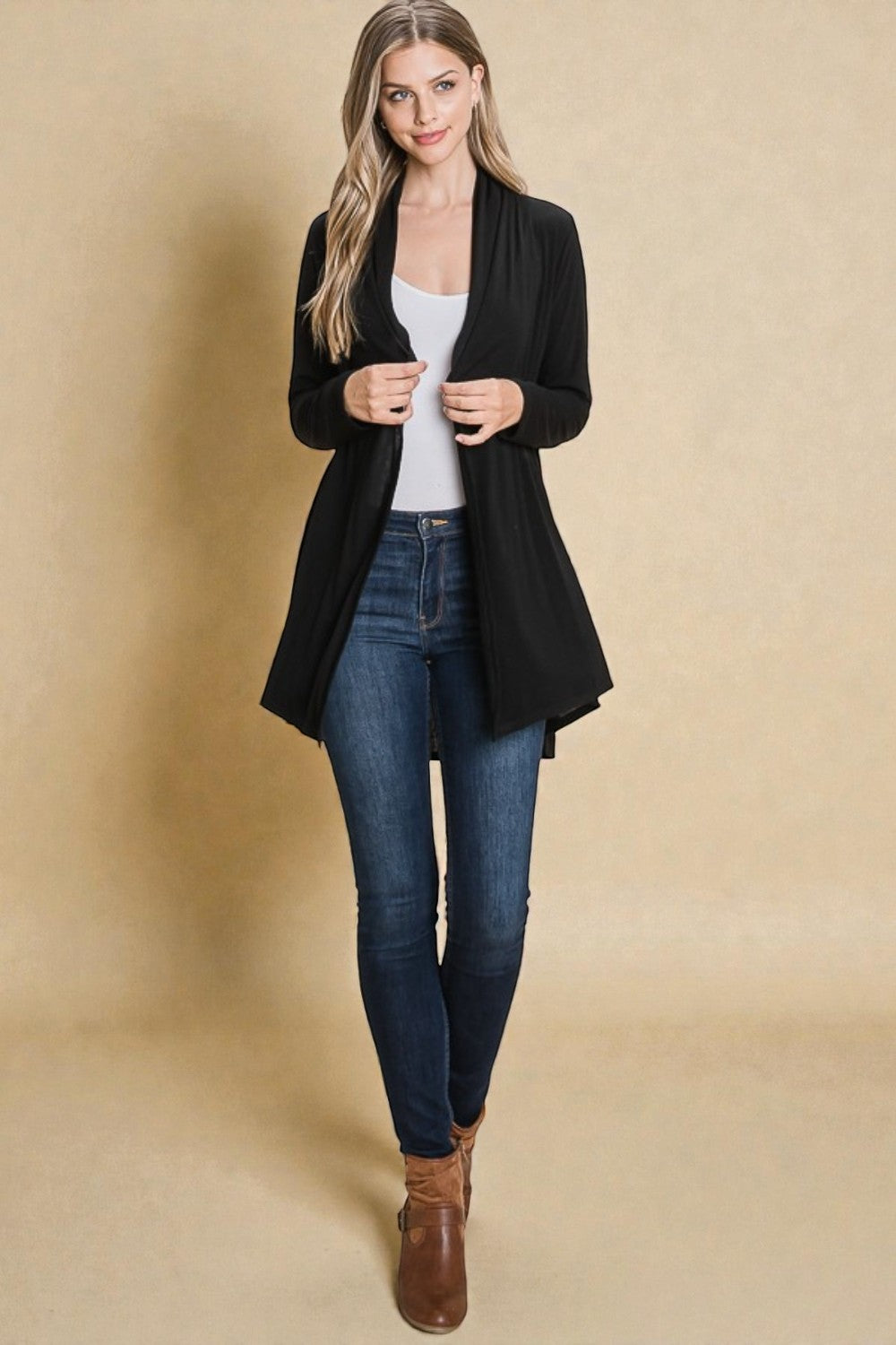 Woman wearing a black cardigan, white top, blue jeans, and brown boots on a beige background