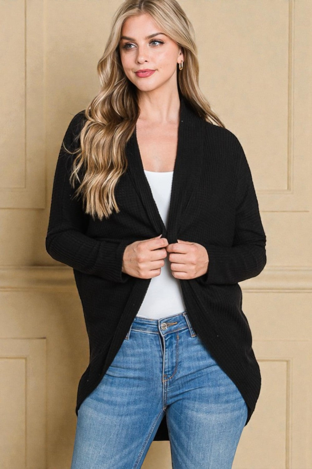Woman wearing a black cardigan over a white top and blue jeans against a beige wall.
