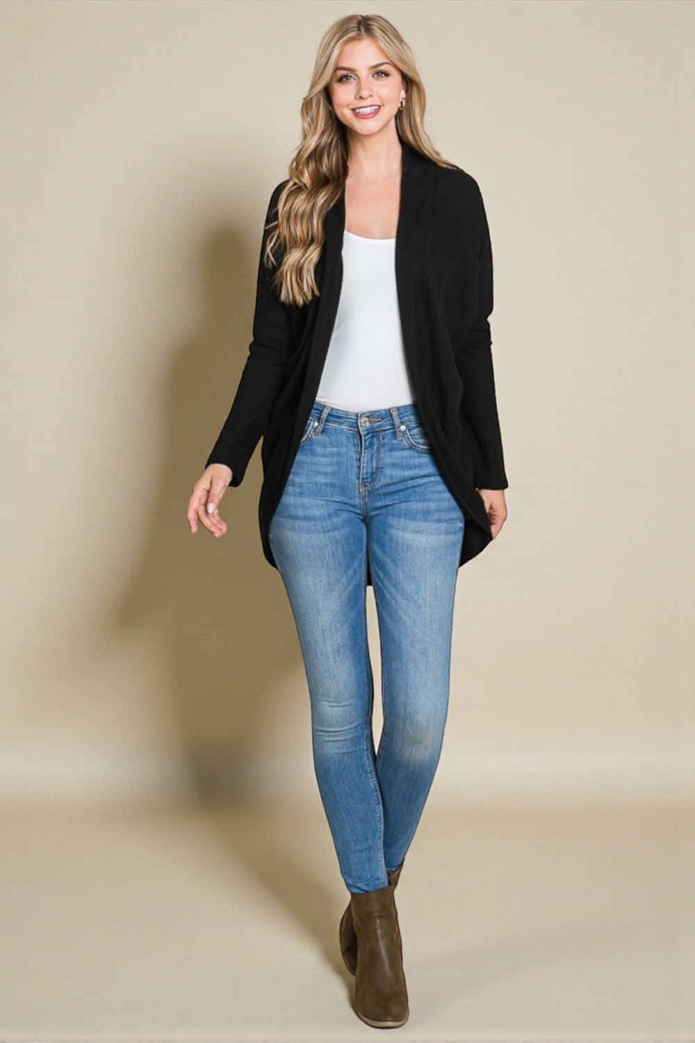 Woman wearing a black cardigan, white top, blue jeans, and brown boots against a beige background
