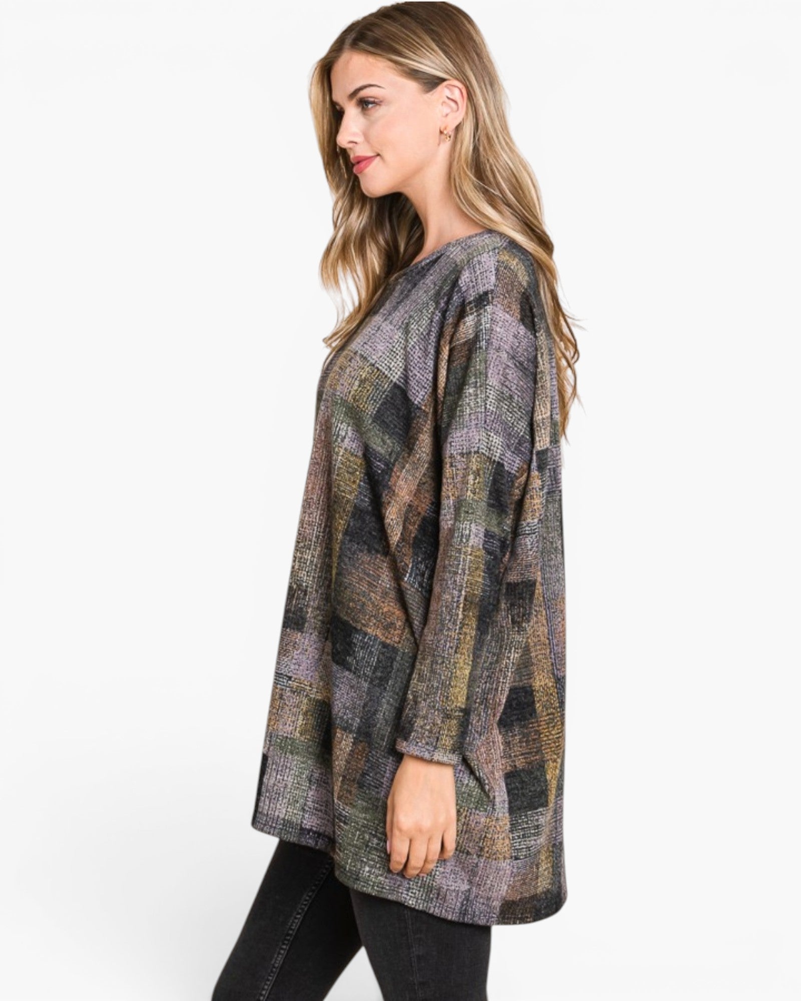 A woman modeling a loose, long-sleeve patchwork plaid tunic top in muted earthy tones of gray, brown, green, and beige, paired with black pants against a plain white studio background.