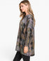 A woman modeling a loose, long-sleeve patchwork plaid tunic top in muted earthy tones of gray, brown, green, and beige, paired with black pants against a plain white studio background.