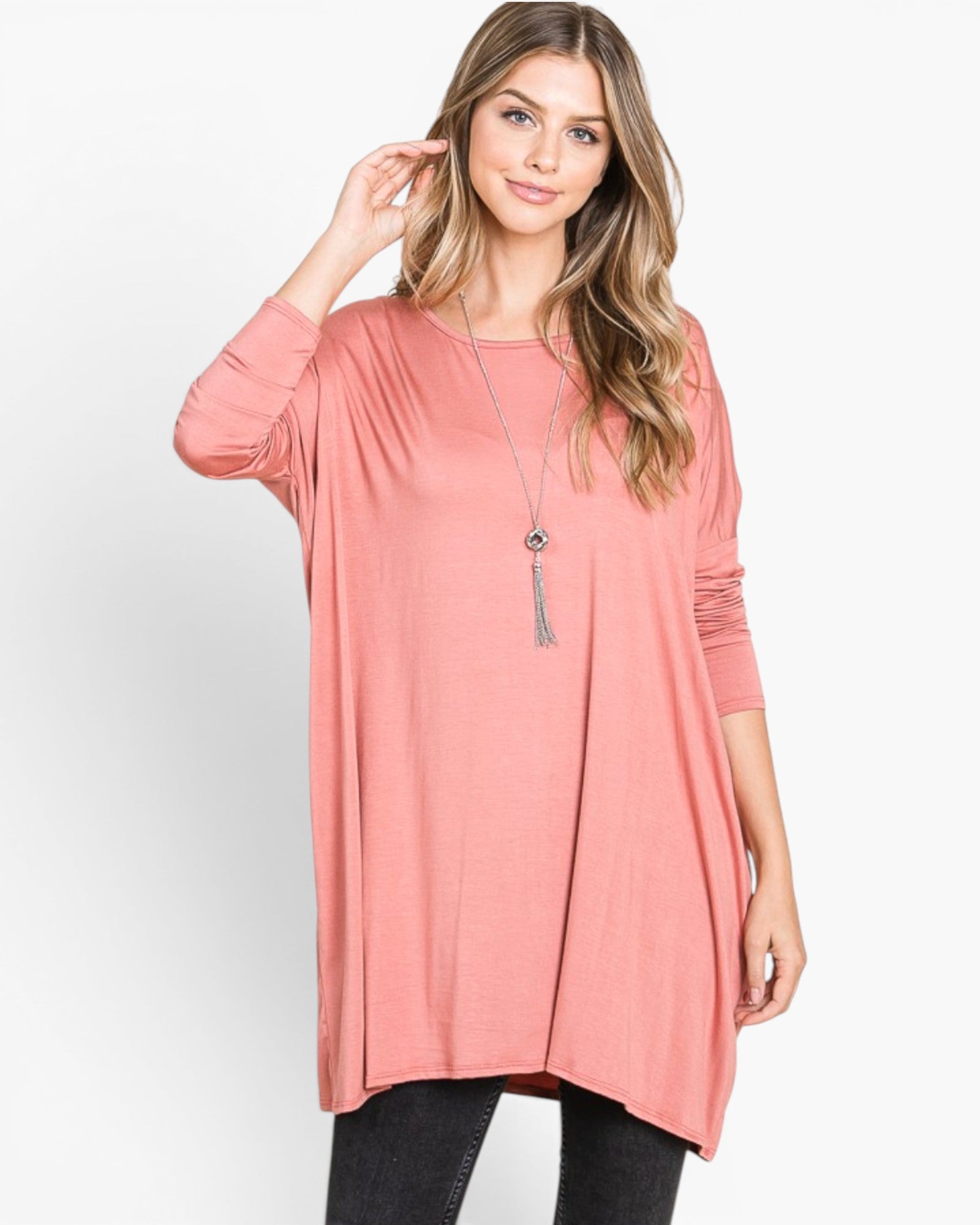 A woman modeling a dusty pink long-sleeve tunic top with a relaxed, flowy silhouette, paired with black pants and a silver tassel necklace, against a plain white studio background.