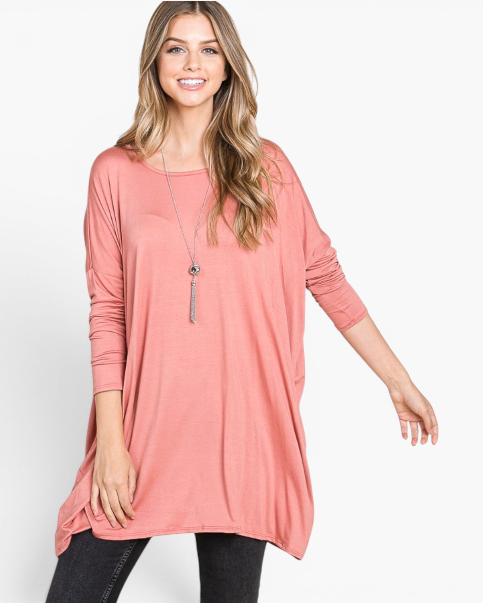 A woman modeling a loose, dusty pink long-sleeve tunic top with a relaxed fit and high-low hem, paired with black pants against a plain white studio background.