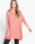 A woman modeling a loose, dusty pink long-sleeve tunic top with a relaxed fit and high-low hem, paired with black pants against a plain white studio background.