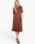 A woman modeling a short-sleeve midi dress with a rust-brown and terracotta mottled pattern, paired with black slide sandals, against a plain white studio background.