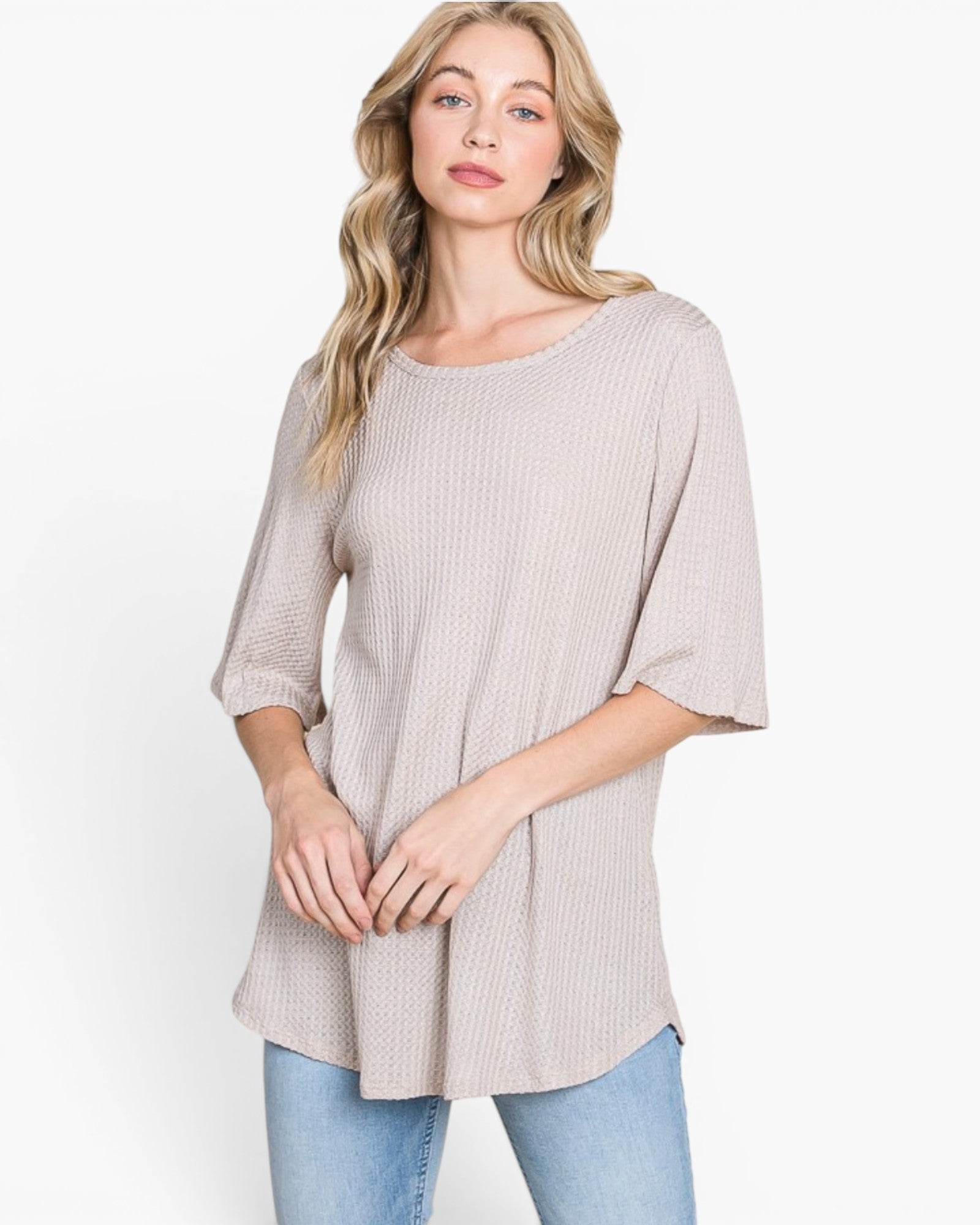 A woman wearing a light beige ribbed knit top with three-quarter sleeves and a relaxed fit, paired with blue jeans, against a plain white studio background.
