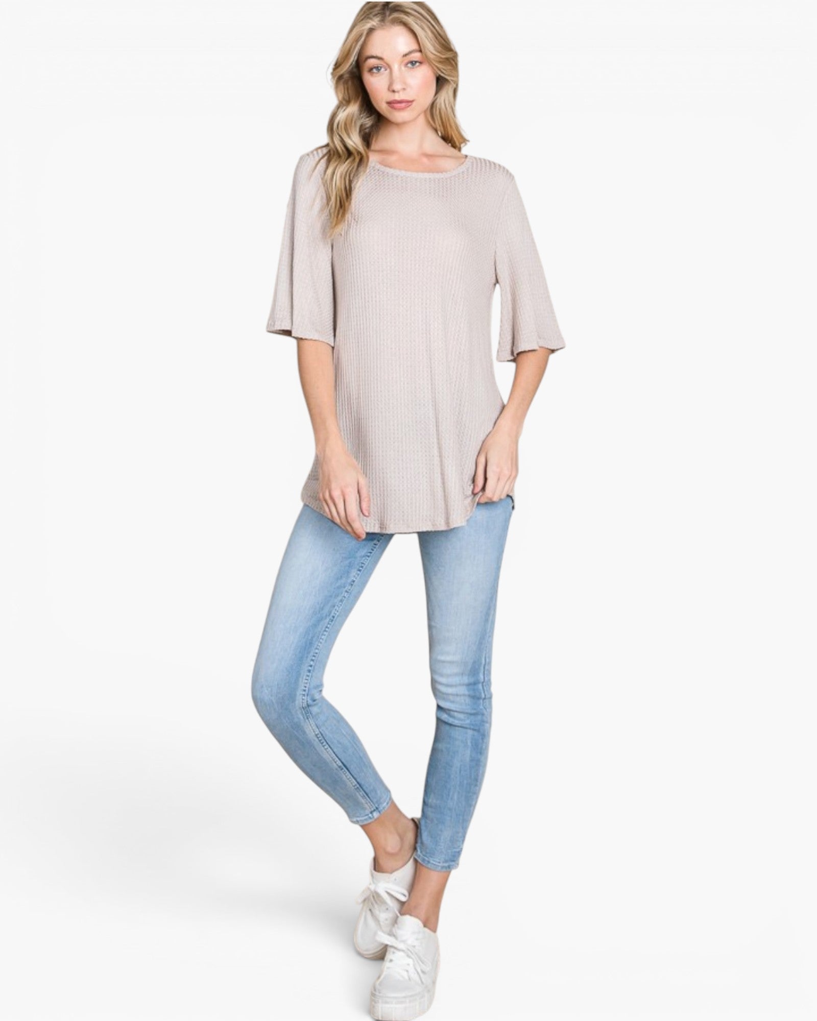 A woman modeling a light beige short-sleeve tunic top with elbow-length flared sleeves, paired with light blue skinny jeans and white sneakers, against a plain white studio background.