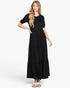 A woman modeling a black tiered maxi dress with short puff sleeves and white slide sandals against a plain white studio background.