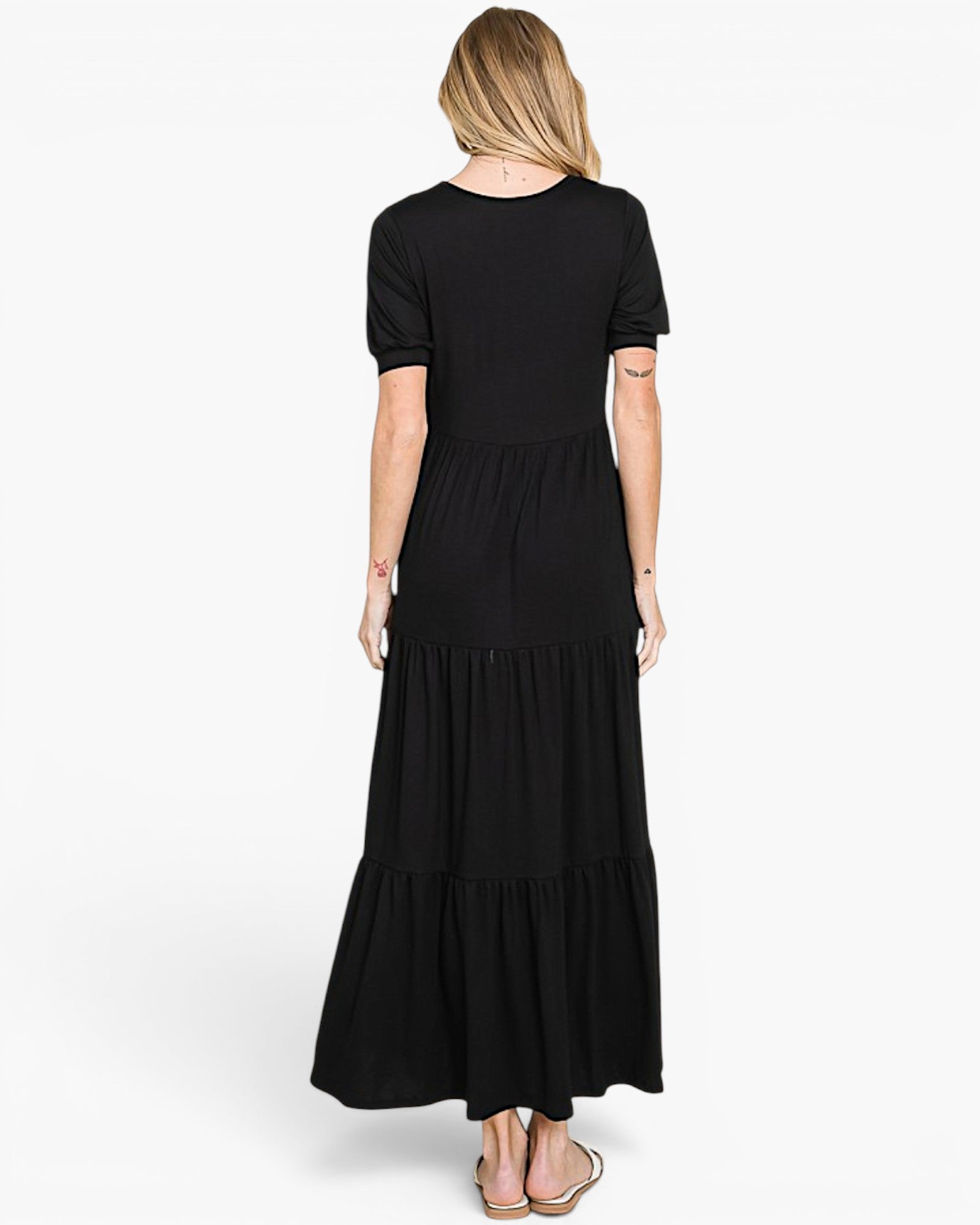 A woman viewed from the back wearing a black short-sleeve maxi dress with a tiered skirt, paired with beige flat sandals, against a plain white studio background.