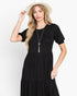 A woman modeling a black tiered midi dress with short puff sleeves and a gathered waist, styled with a long silver pendant necklace and a light beige wide-brim hat against a plain white background.