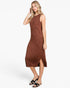 A woman modeling a sleeveless rust-brown midi dress with a subtle textured pattern and a side slit, paired with black slide sandals, against a plain white studio background.