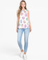 A woman modeling a sleeveless white top with a colorful smiley face pattern, paired with light blue skinny jeans and white sneakers, against a plain white background.