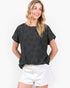 A woman wearing a black short-sleeve t-shirt with a subtle tonal floral pattern, paired with white shorts, against a plain white background.