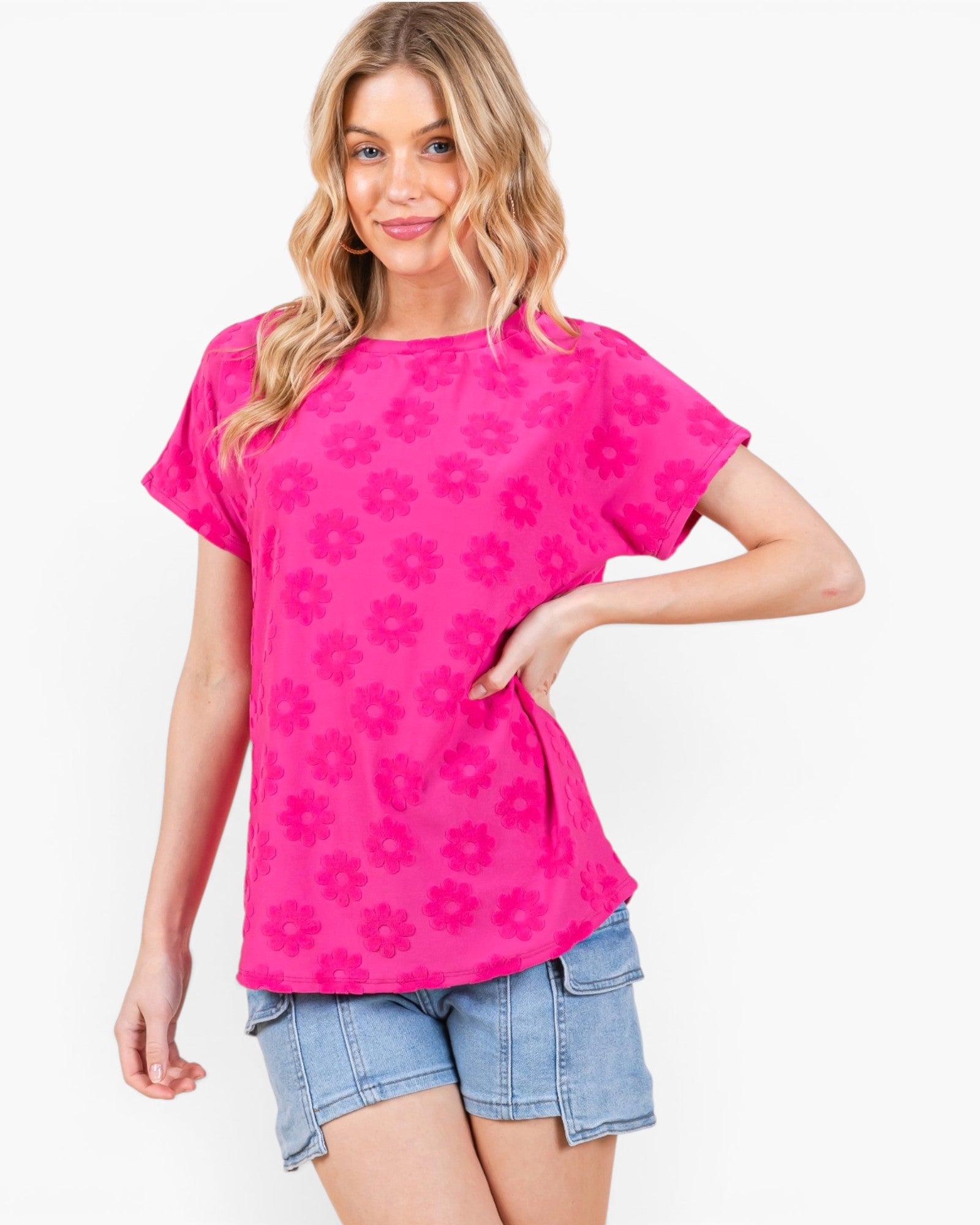 A woman modeling a bright pink short-sleeve top with a tonal floral pattern, paired with denim shorts, against a plain white background.