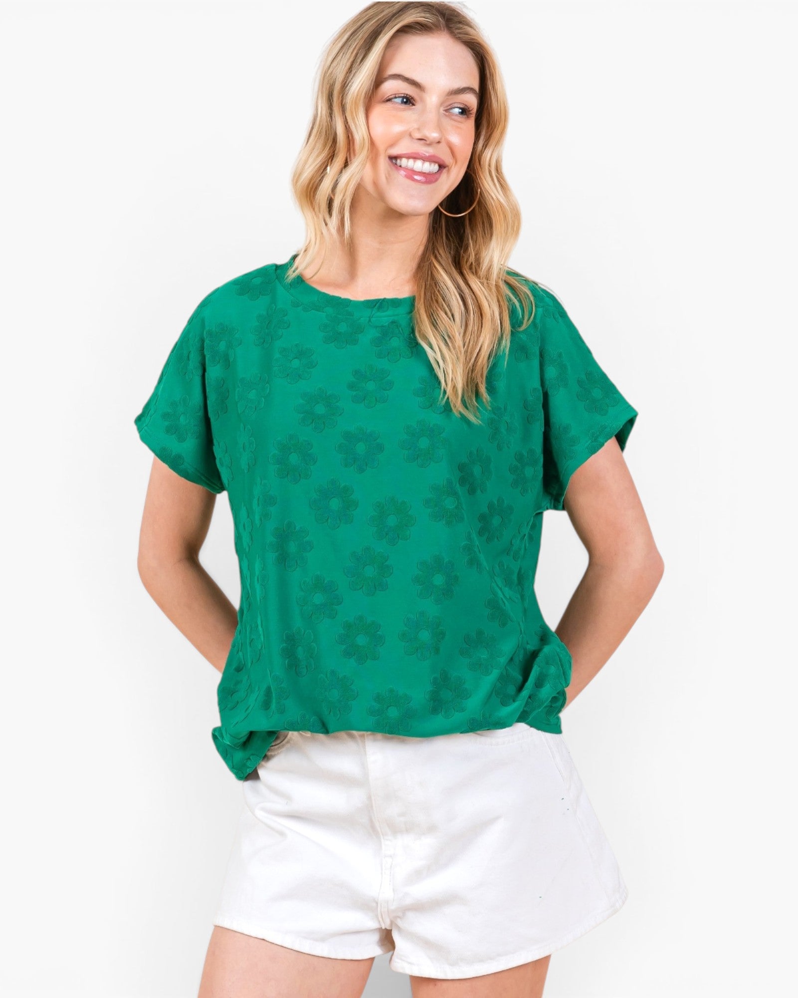 A woman wearing a green short-sleeve top with a tonal floral pattern and white shorts, standing against a plain white background.