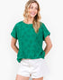 A woman wearing a green short-sleeve top with a tonal floral pattern and white shorts, standing against a plain white background.