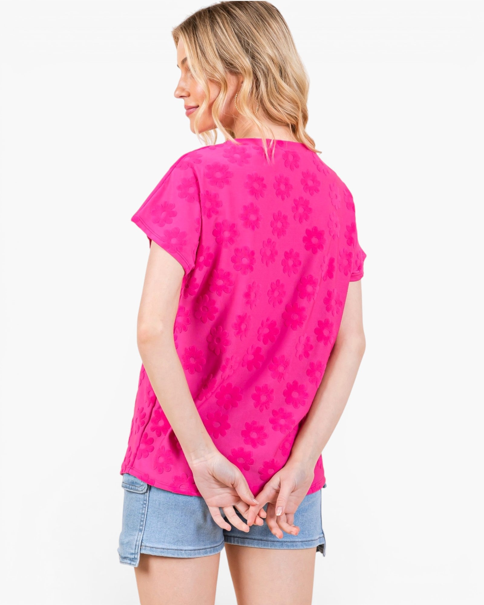 A woman shown from the back wearing a bright pink short-sleeve top with a tonal floral pattern and light blue denim shorts against a plain white background.