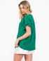A woman wearing a green short-sleeve top and white shorts, photographed against a plain light gray studio background.