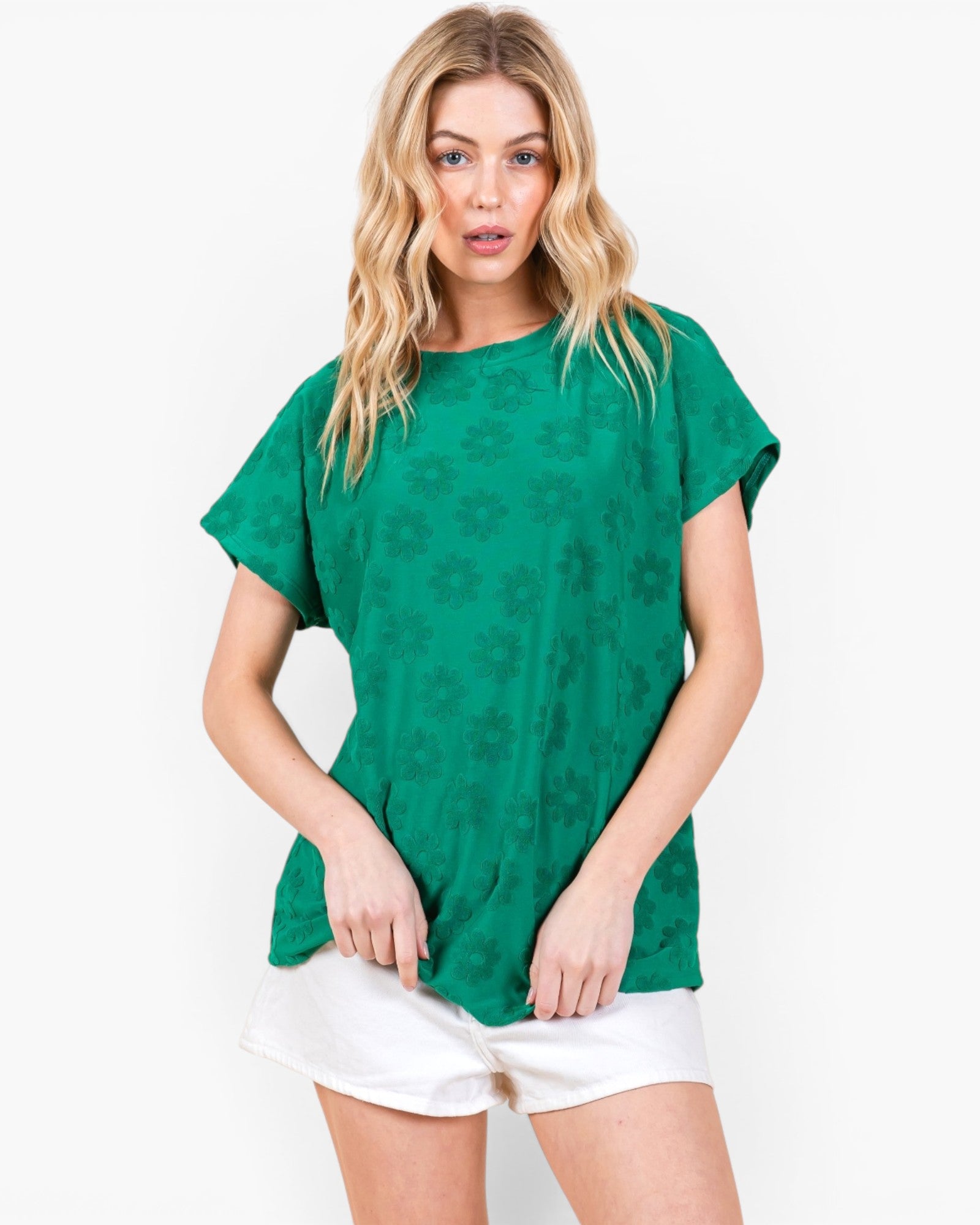 A woman modeling a green short-sleeve t-shirt with a tonal floral pattern, paired with white shorts, against a plain white background.