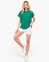 A woman modeling a green short-sleeve t-shirt with a tonal floral pattern, paired with white shorts and white sneakers, against a plain white background.