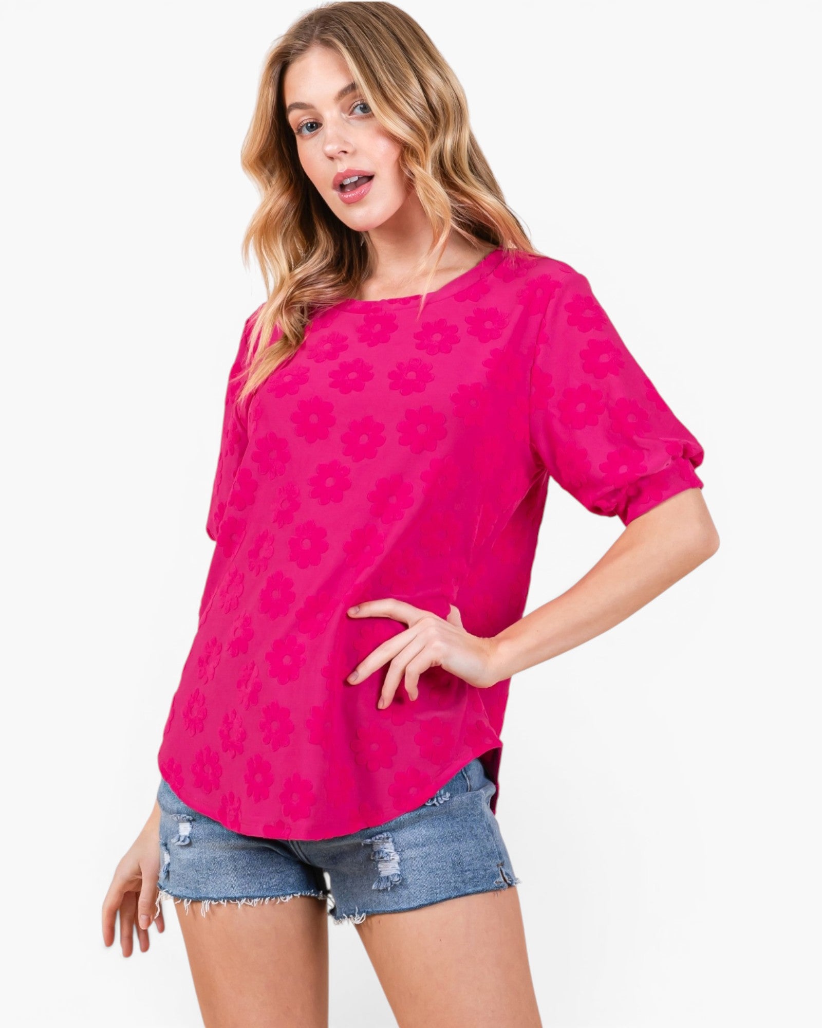 A woman modeling a bright pink short-sleeve top with a tonal floral pattern, paired with distressed denim shorts, against a plain white studio background.