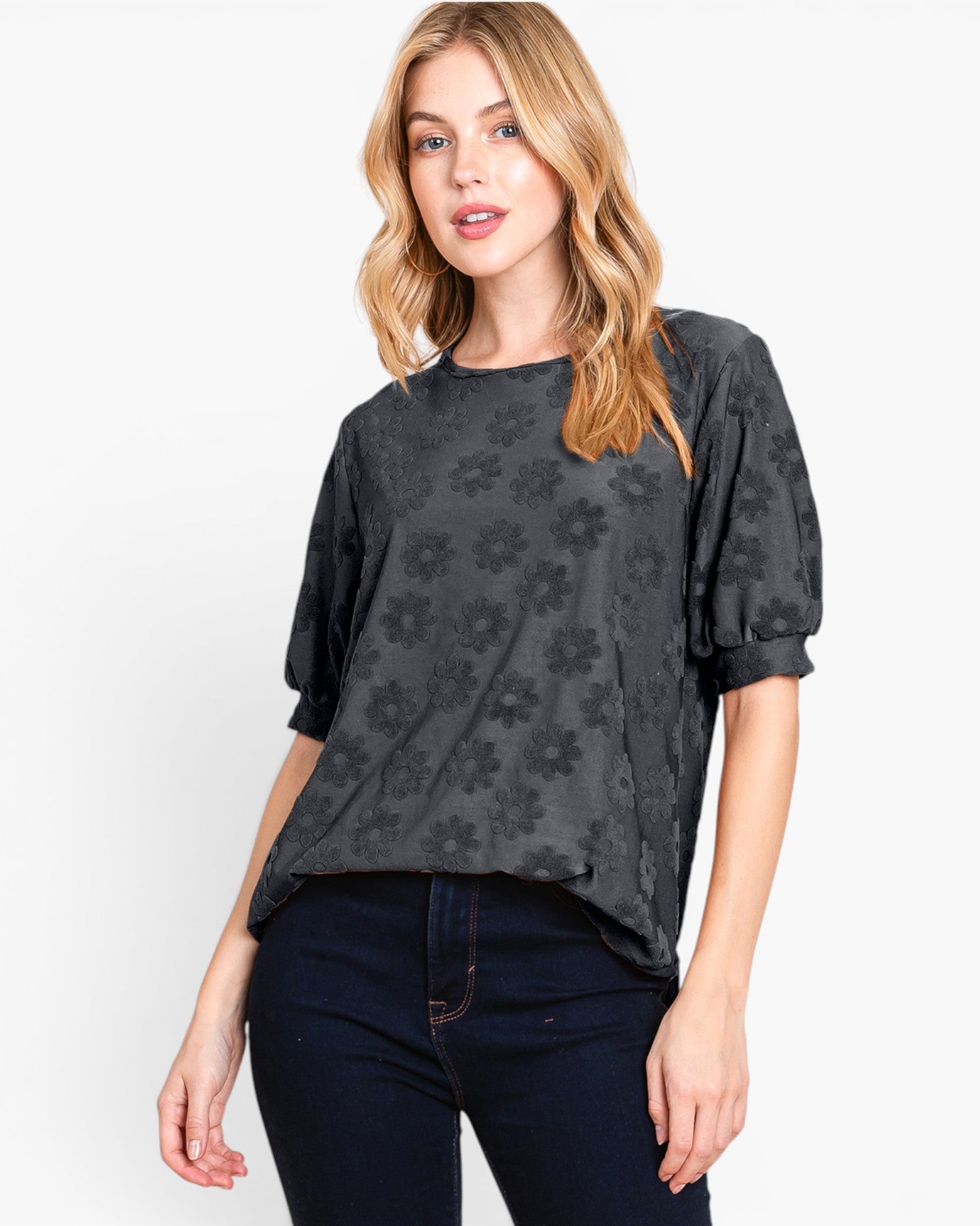 A woman modeling a dark gray short-sleeve top with a tonal floral pattern, paired with dark blue jeans, against a plain white background.