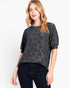A woman modeling a dark gray short-sleeve top with a tonal floral pattern, paired with dark blue jeans, against a plain white background.
