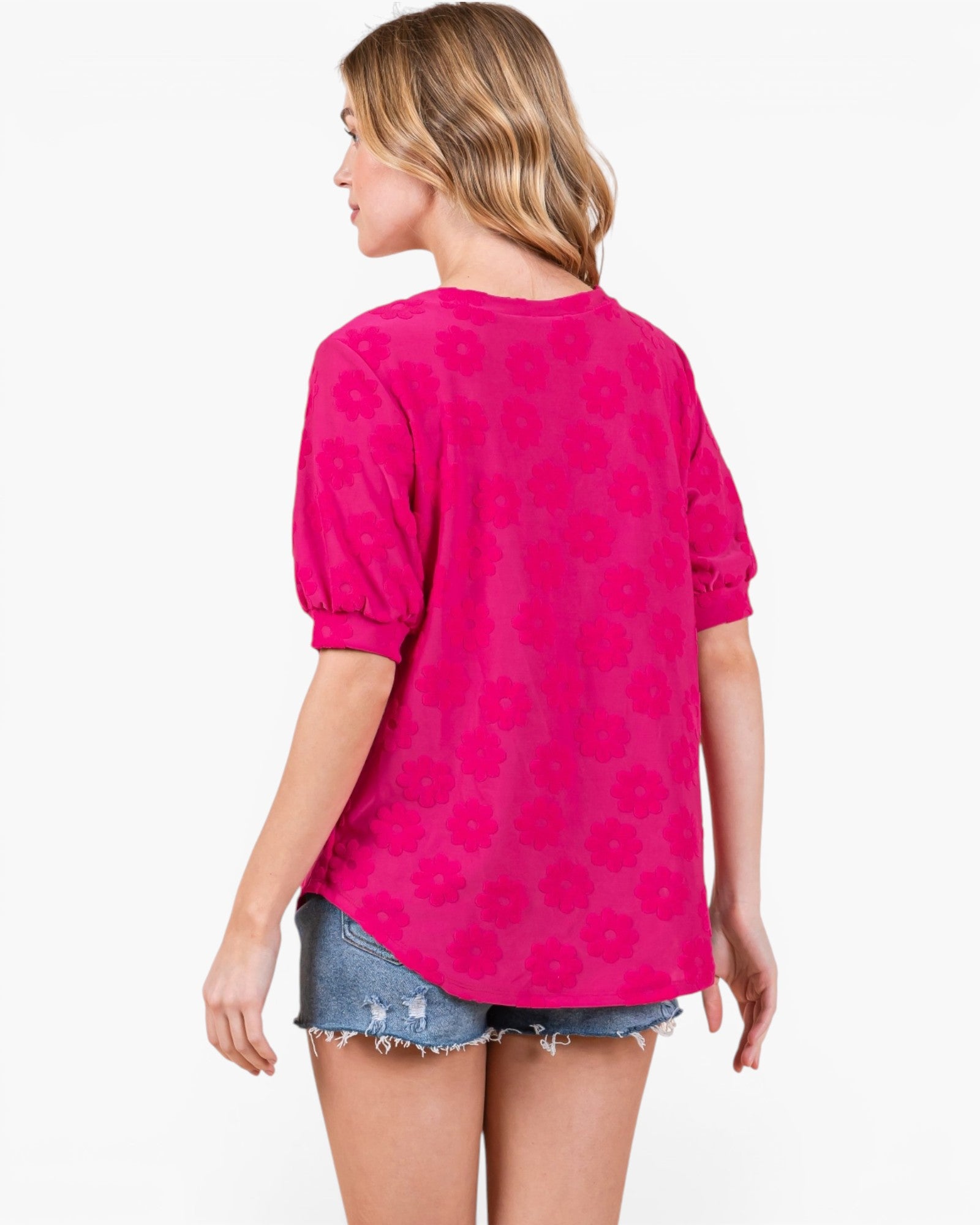 A woman shown from the back wearing a bright pink short-sleeve top with a tonal floral pattern and denim shorts against a plain white background.