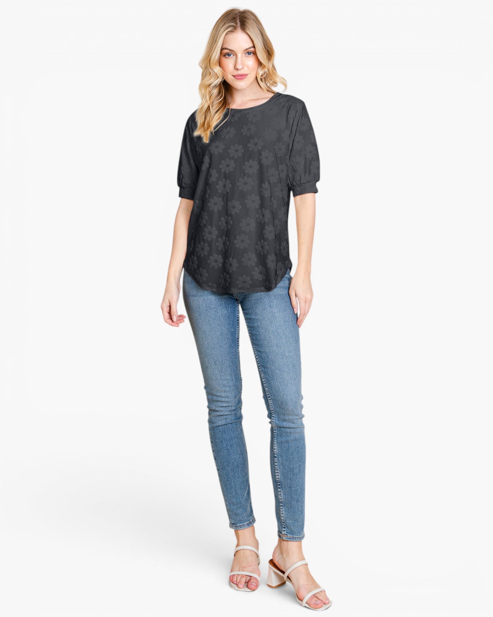 A woman modeling a short-sleeve black top with a subtle tonal floral pattern, paired with blue skinny jeans and white heeled sandals, against a plain white studio background.