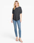 A woman modeling a short-sleeve black top with a subtle tonal floral pattern, paired with blue skinny jeans and white heeled sandals, against a plain white studio background.