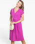 A woman modeling a bright magenta short-sleeve V-neck dress with a button placket, holding a cream-colored crossbody bag against a plain white studio background.