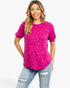 A woman modeling a bright fuchsia short-sleeve top with a tonal textured pattern, paired with distressed light-wash blue jeans, against a plain white studio background.