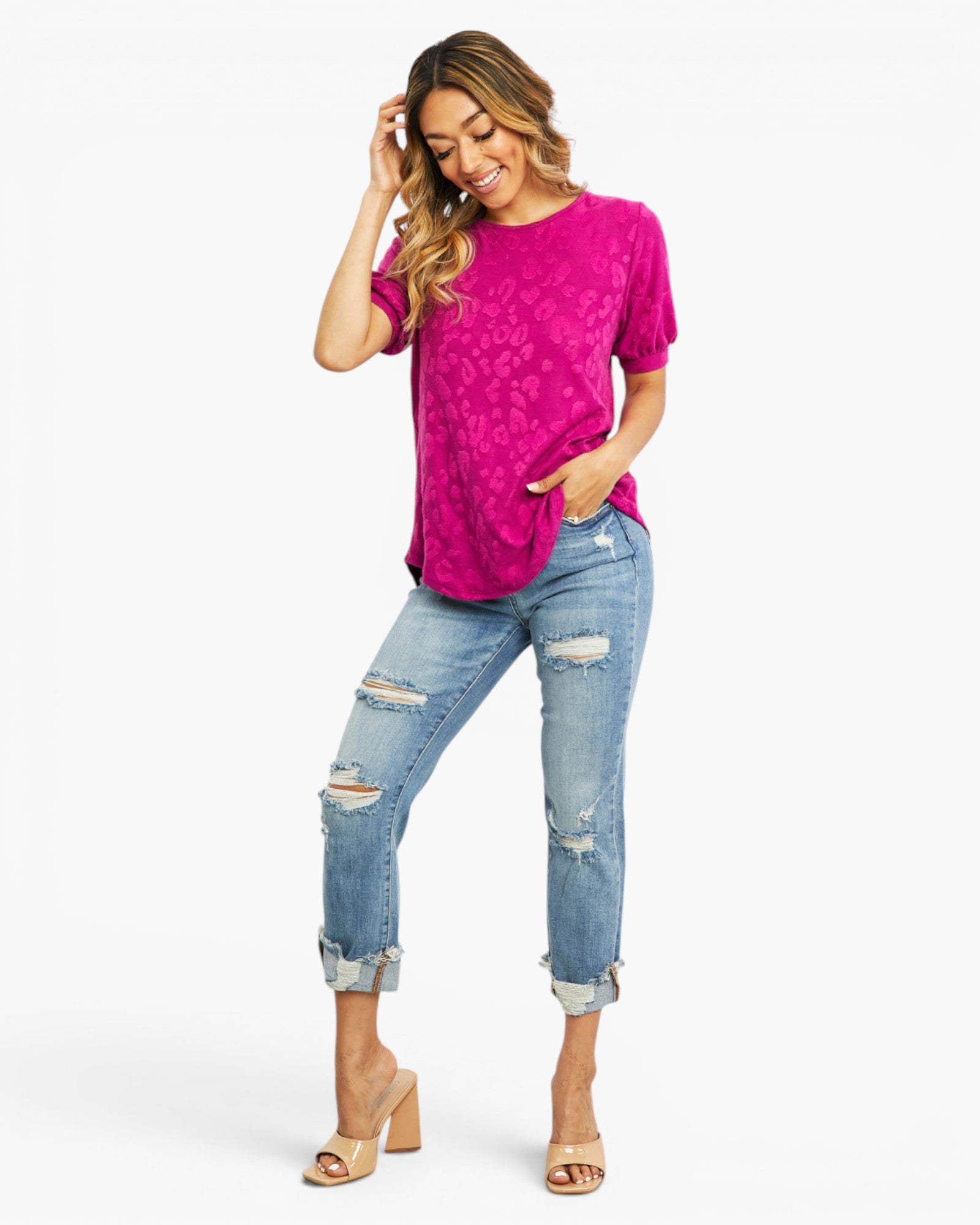 A woman modeling a bright pink short-sleeve t-shirt with a subtle tonal floral pattern, paired with light-wash distressed cuffed jeans and beige heeled sandals, against a plain white studio background.